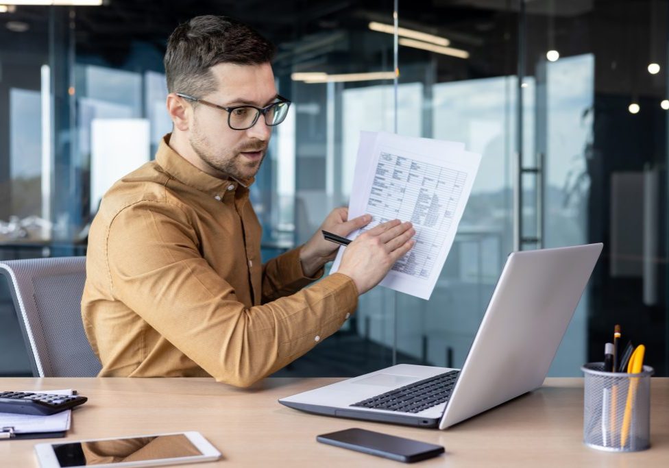 A young male businessman conducts online business training, a conference, sitting in the office at the table. Shows documents, a project on the laptop monitor.
