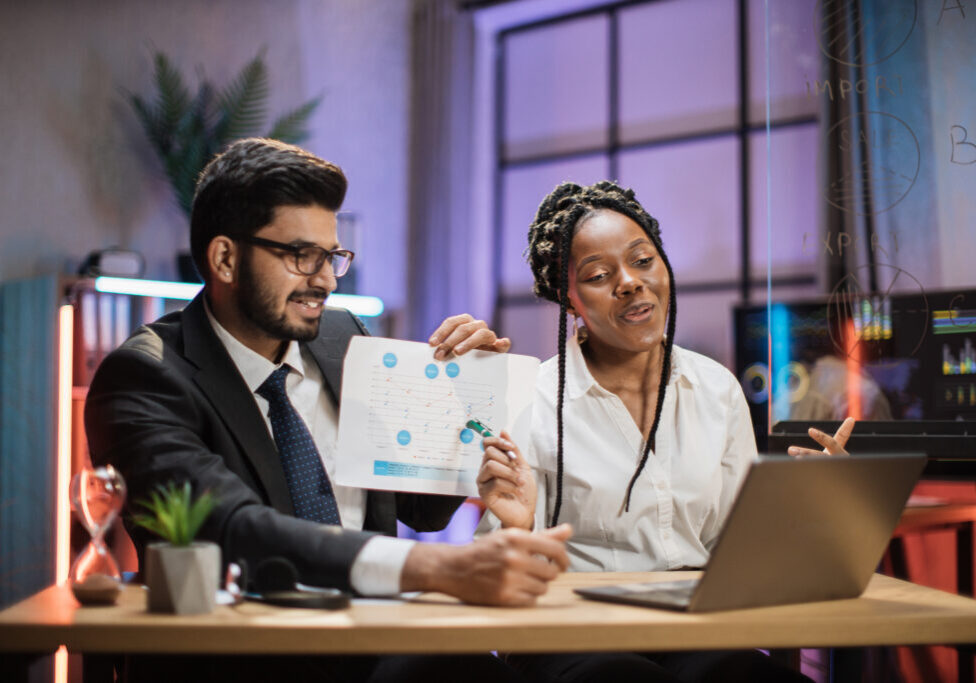 Two multiracial brokers, handsome arab bearded office manager and african businesswoman during online video call with coworkers and explaining report with diagram. Office working at night concept.