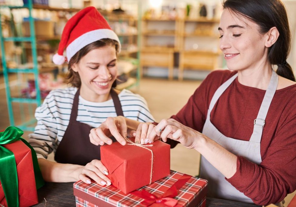 Portrait of two smiling young women wrapping gift boxes for Christmas sitting at table in store
